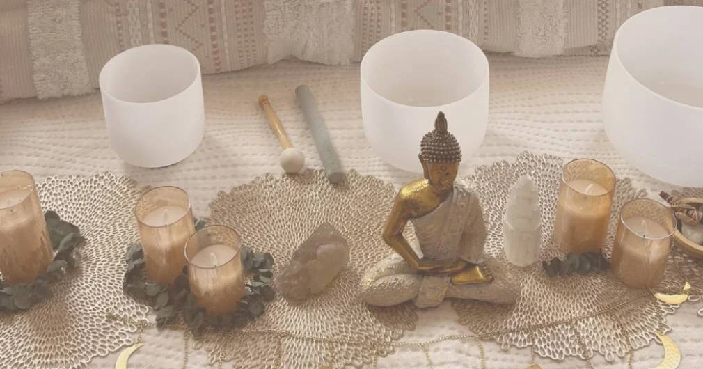 Crystal singing bowls, glowing candles, two crystals and a golden Buddha statue arranged on a cream cloth