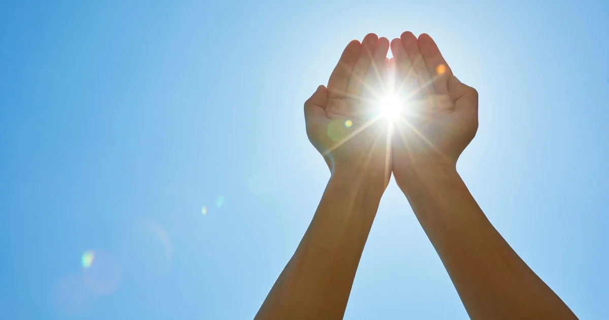 Two hands cupped together and raised against a clear blue sky, holding a bright burst of sunlight
