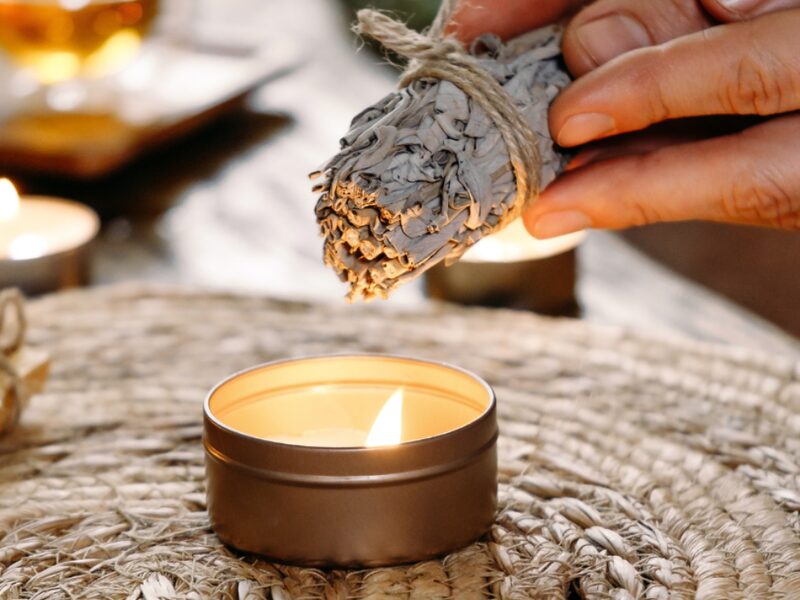 A hand holds a bundle of dried sage above a lit tin candle, with more candles glowing behind it
