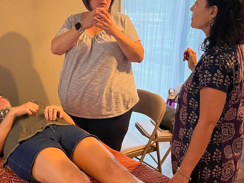 Two women standing over a person who lies on a treatment table during a hands-on class