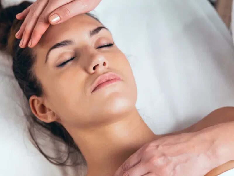 A woman with closed eyes lies on a white table while a practitioner rests a hand on her forehead