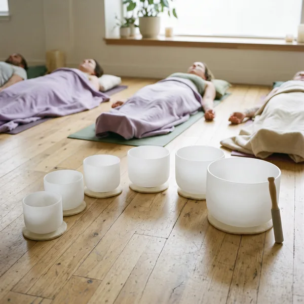Frosted white crystal singing bowls in a row on a wooden floor, with people resting under blankets on mats behind them